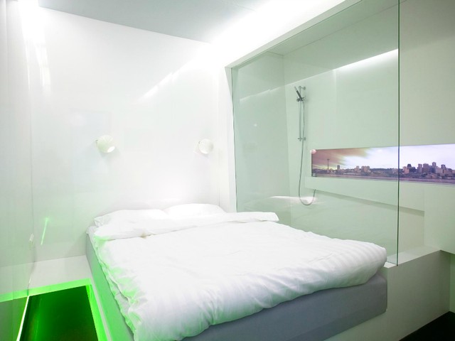 A white hotel room with glass walls, a city panorama and a vivid green light beneath the bed.